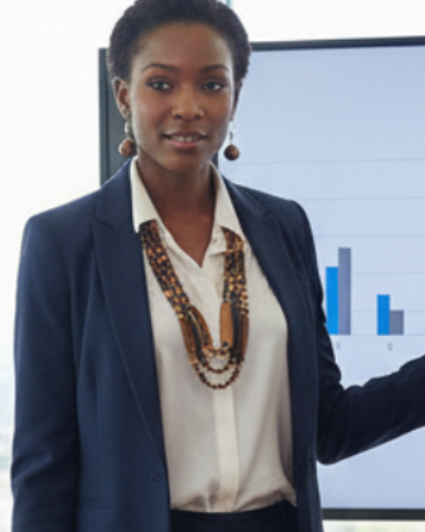 A professional woman in a navy blazer and white blouse stands at a conference table in a modern office, gesturing toward a large screen showing a blue bar chart. Several men seated around the table are taking notes, with a wood conference table and large windows in the background.