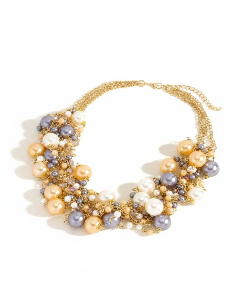 A gold chain statement necklace adorned with faux pearls in cream, lavender, and gold, arranged in a tiered collar style on a light beige background.