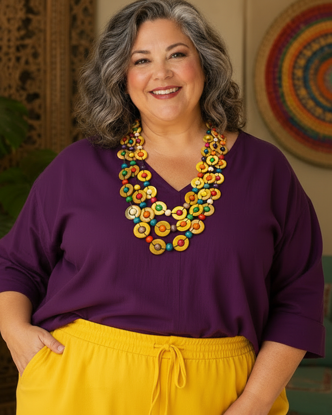 A smiling woman with gray hair is wearing a bright yellow V-neck top and purple drawstring pants, accessorized with a chunky multi-colored beaded necklace. She is posed indoors against a warm-toned background with plants and woven decor.
