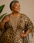 A woman with short gray hair models a leopard print dress with wide draped sleeves and a beaded fringe neckline. She is wearing gold-toned jewelry and standing in front of a beige wall with a green potted plant on the left.