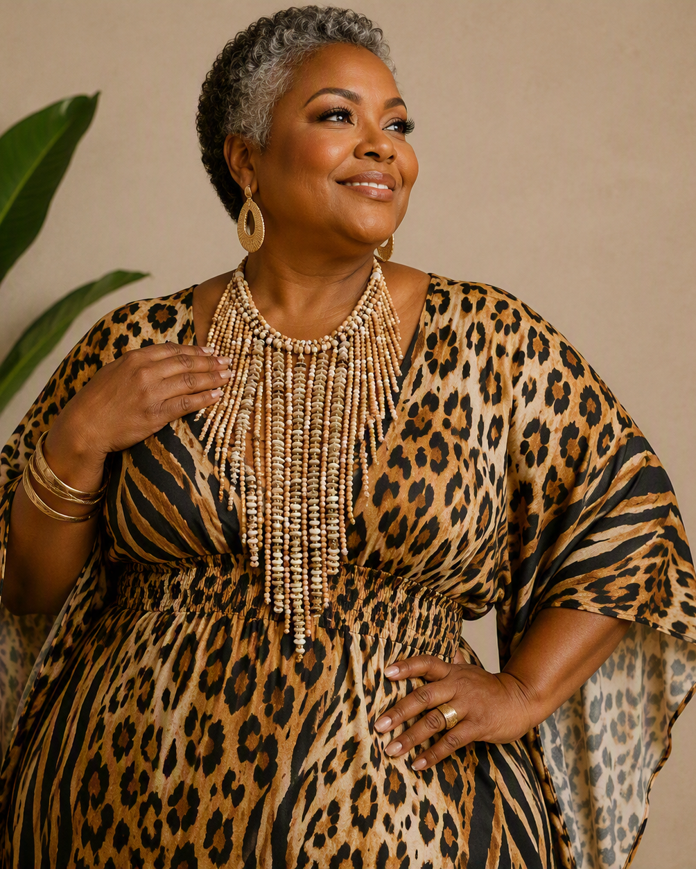 A woman with short gray hair models a leopard print dress with wide draped sleeves and a beaded fringe neckline. She is wearing gold-toned jewelry and standing in front of a beige wall with a green potted plant on the left.