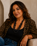 A woman with wavy brown hair sits in a beige armchair wearing a leopard-print cardigan over a black top and blue jeans. She is accessorized with a chunky black beaded necklace and gold hoop earrings, with a rustic brown vase of dried grass in the background.